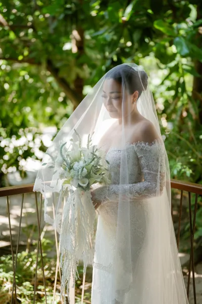 Bridal portrait of a bride under veil, wearing a lace wedding dress, softly lit amid garden greenery beside a wood railing