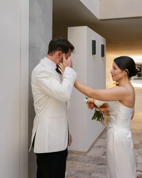 First look moment as bride touches groom’s face, peach and white bouquet in hand, outside a modern venue with concrete columns
