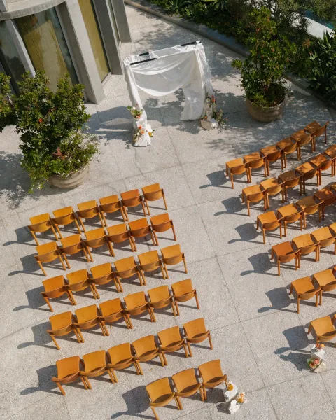 Ceremony setup with white draped arch and floral arrangements, wooden chairs on a stone patio beside a modern courtyard building exterior