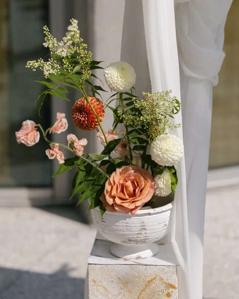 Wedding floral arrangement of peach rose and white dahlias in a ceramic urn on a stone pedestal with soft white drapery backdrop
