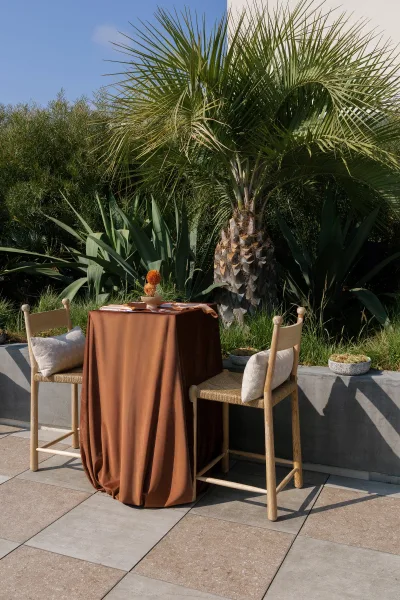 Reception tablescape with an outdoor sweetheart table for two, brown linen, wooden chairs, simple place settings, and succulents on a stone patio with palm and desert greenery