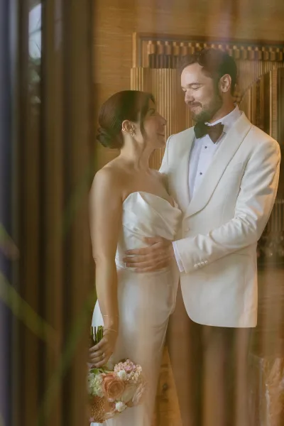 Couple portrait of bride looking at groom, wearing strapless satin wedding dress and holding bouquet, posed by a wood-paneled wall