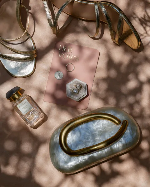 Wedding flat lay of wedding rings in a box beside a vow book, gold strappy heels and perfume on blush fabric with shadows