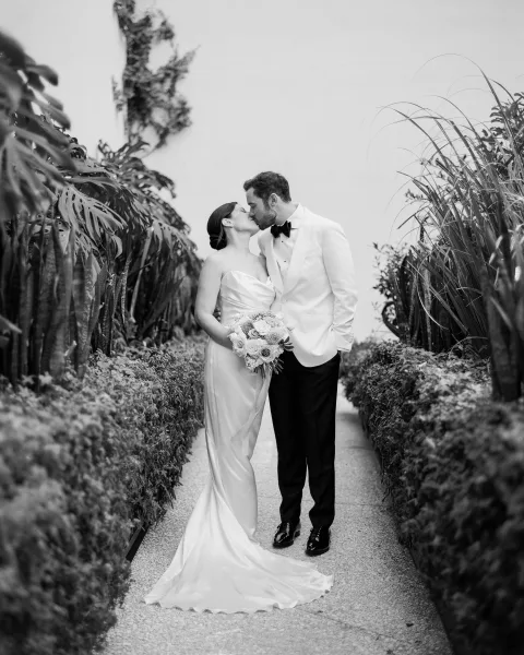 Wedding kiss portrait of the bride and groom kissing on a garden path, she holds a bridal bouquet beside tropical plants and hedges