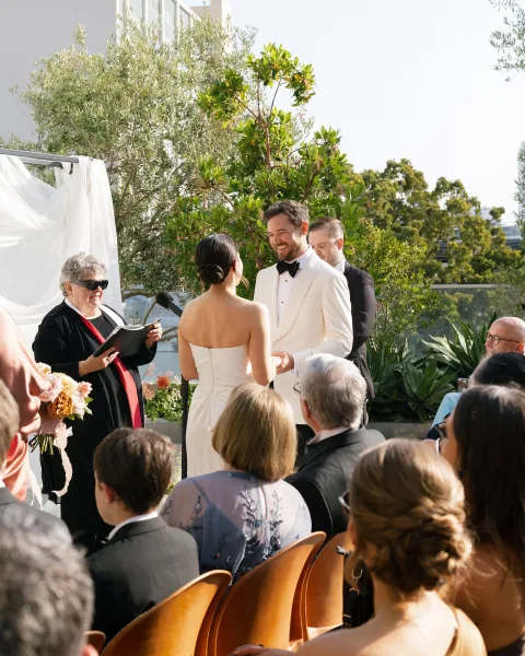 Wedding vows shared as bride and groom hold hands at an outdoor wedding ceremony on a terrace, officiant at mic under white drape backdrop