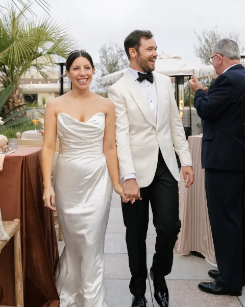 Newlywed couple holding hands as they walk into an outdoor reception, bride in strapless satin gown and groom in white jacket under palm trees