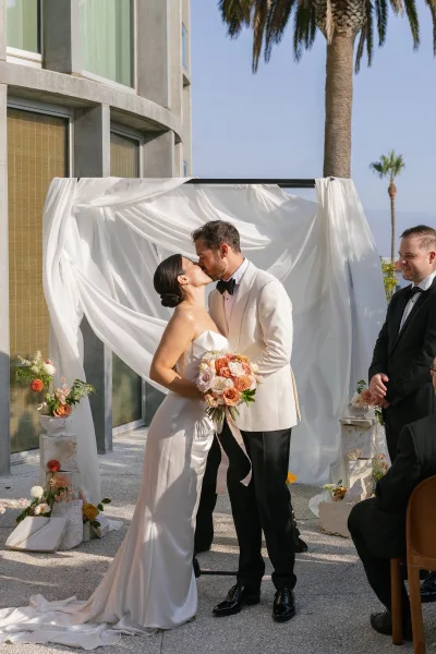 Wedding kiss at the first kiss ceremony under a draped arch, bride in strapless satin gown holding bouquet on a sunny courtyard patio
