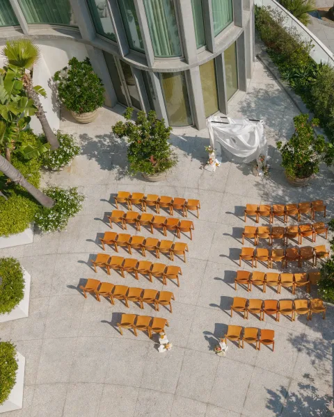 Outdoor ceremony setup with wood ceremony chairs facing a white draped arch, minimal aisle florals on a stone patio by a modern venue