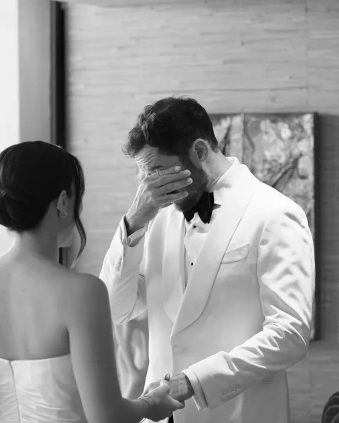 First look moment as groom in a white tuxedo jacket wipes tears while holding hands with the bride in soft indoor window light