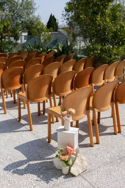 Ceremony seating with tan leather chairs lined on a stone terrace aisle, bud vases and peach-blush roses on stone plinths