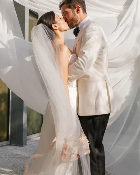 Wedding kiss portrait of bride and groom kissing under a flowing veil, with bouquet and sheer draped fabric backdrop by windows