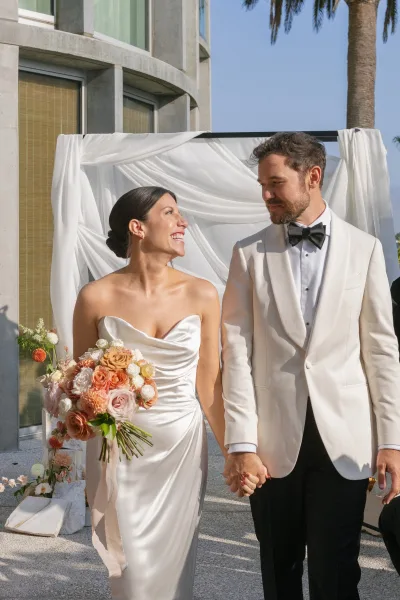 Couple portrait of bride and groom holding hands, bride with bouquet and strapless dress, groom in white tux, by modern facade and palm tree