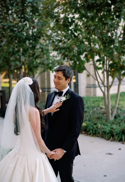 Couple portrait of a bride and groom holding hands, the bride touching his white rose boutonniere, framed by garden trees and a building facade.