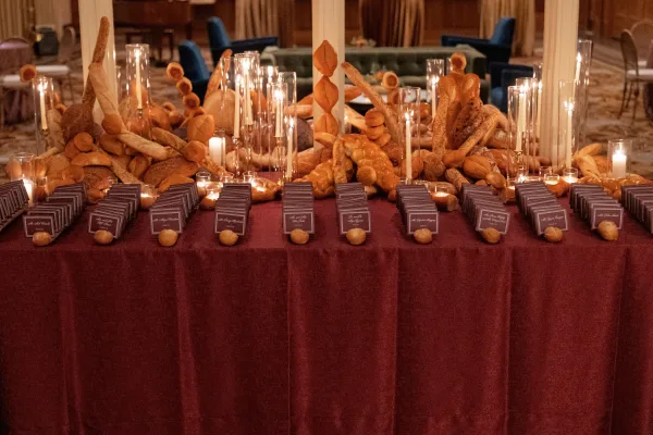 Escort card display featuring edible escort cards tucked into bread loaves and rolls with taper candles on burgundy linen in a paneled ballroom.