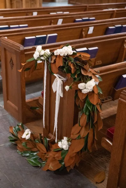 Ceremony aisle decor with church pew flowers featuring white roses at pew ends, flanking wooden pews with hymnals on a cool stone floor.