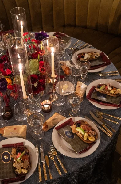 Reception tablescape set as a candlelit wedding table, anchored by a red floral centerpiece against velvet banquette seating indoors.