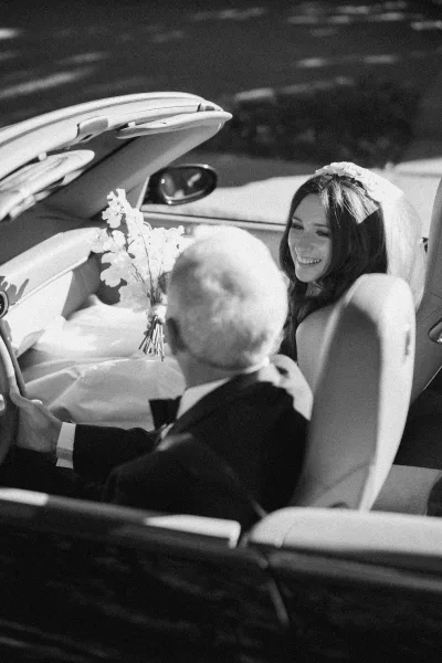 Bride in getaway car beams from a classic convertible, ready for a joyful wedding send-off along a sunlit street.