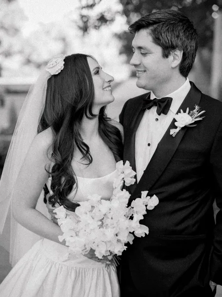 Couple portrait of a bride and groom smiling, the bride holding a simple bridal bouquet, set in a leafy garden with trees and foliage.