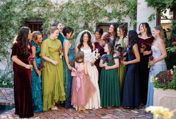 Bride and bridesmaids in mismatched bridesmaid dresses gather as the bride holds a white bridal bouquet in a vine-covered courtyard.