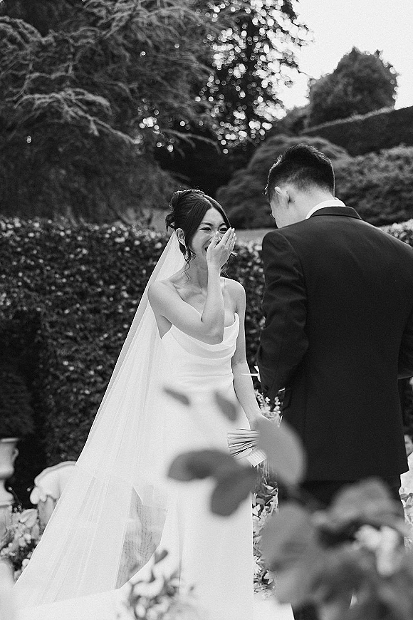 First look moment as bride in a strapless dress and long veil laughs covering her face while groom reacts in a garden setting