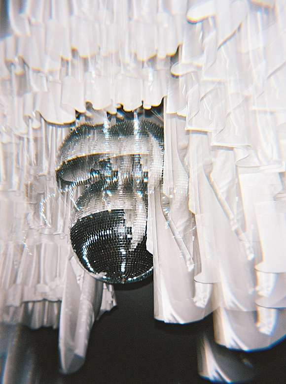 Disco ball decor with a wedding disco ball hanging amid ruffled white fabric drapery against a white curtain backdrop indoors