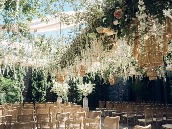 Ceremony setup with outdoor ceremony decor featuring hanging florals, set against a brick wall and evergreen trees under a blue sky.