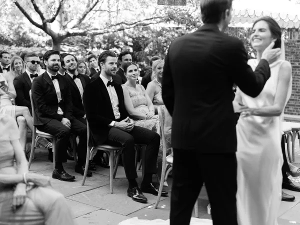 Ceremony moment during the wedding vows moment as the bride laughs and the groom caresses her, her veil lifting on a stone patio beneath garden trees.
