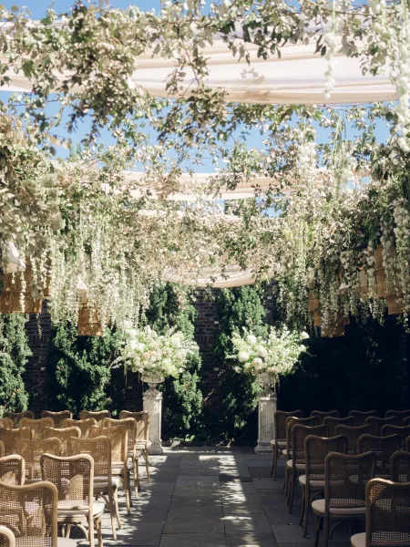 Ceremony setup highlighting outdoor ceremony decor with hanging florals along a stone patio framed by garden trees and a warm brick wall.