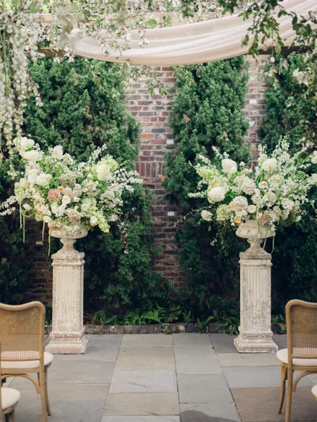 Ceremony altar with outdoor ceremony decor framed by draped fabric against a brick wall and evergreen trees on a stone patio.