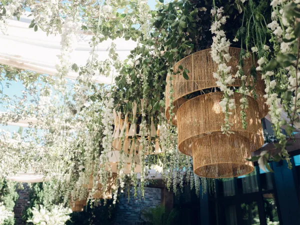 Hanging floral installation featuring greenery ceiling decor with rattan chandeliers, set against a blue sky and brick wall with garden trees.