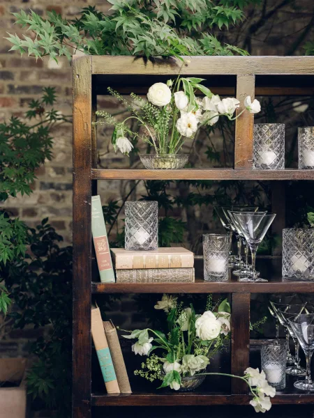 Wedding bar decor with elegant bar back styling featuring crystal glassware against a brick wall with leafy greenery.
