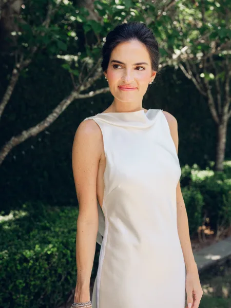 Bridal portrait showcasing a minimalist bridal look in a sleek high-neck sleeveless ivory gown with stud earrings amid garden trees and hedges.