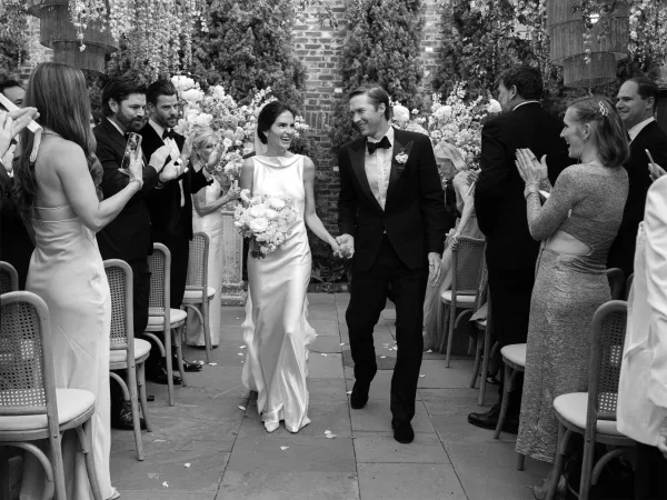 Ceremony recessional as the bride and groom exit, smiling hand in hand past aisle petals, with a brick wall and garden greenery behind.