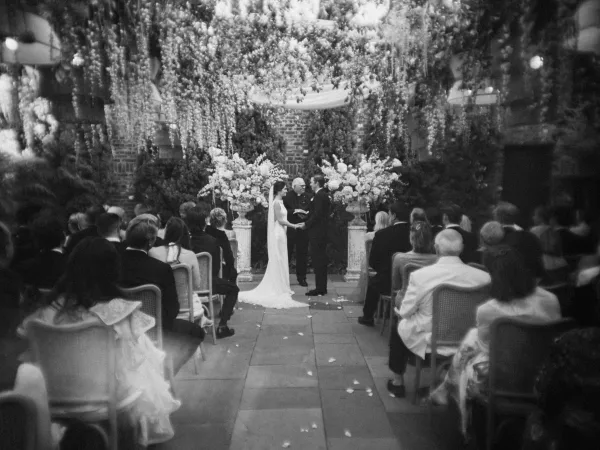 Wedding vows with a couple exchanging vows beneath hanging florals, framed by a brick wall and lush greenery over a stone floor.