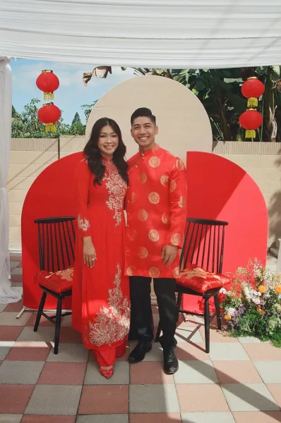 Couple portrait in red traditional wedding outfits, standing by red lanterns and round backdrop under a white canopy on an outdoor patio