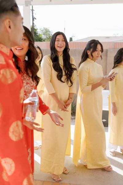 Bridesmaid group of ao dai bridesmaids in yellow dresses, chatting with phones and gold jewelry on a sunny patio under a canopy