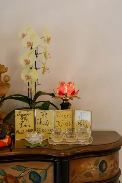 Wedding altar table with orchid plant and lotus candle lamp, framed name cards and tea lights arranged on a wooden cabinet against a plain wall