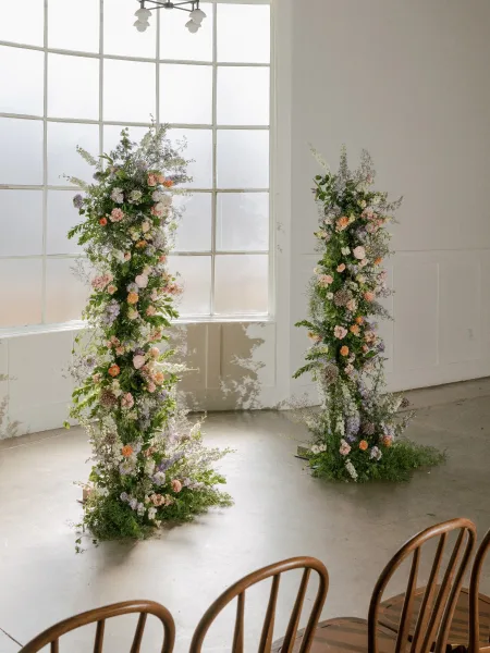 Wedding ceremony arch with floral ceremony pillars in pastel blooms and greenery, set indoors by large grid windows and white walls