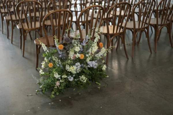 Ceremony aisle flowers in a ground floral arrangement of wildflowers and greenery with orange, white, and lavender blooms beside wood chairs indoors