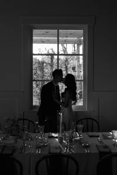Wedding kiss portrait of bride and groom silhouette by a large window, with candlelit reception table settings and greenery outside