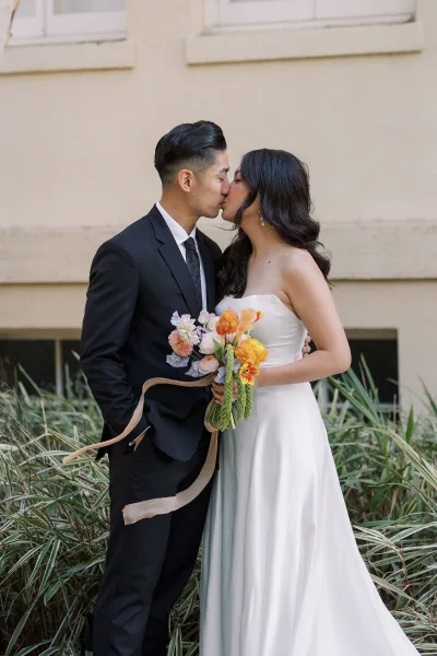 Wedding kiss portrait of bride and groom kissing in side profile, bride in strapless dress holding bouquet with ribbon by stucco wall greenery
