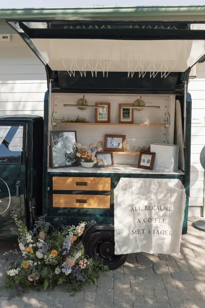 Wedding welcome table with wedding welcome sign, framed photos and floral arrangement beside a mobile bar trailer on stone pavers