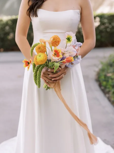 Bridal bouquet of colorful wedding bouquet tulips with long ribbon wrap, held by a bride in a strapless dress on an outdoor hedge walkway