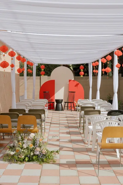 Ceremony setup with a white draped canopy and red hanging lanterns above black chairs, semicircle backdrop panels, and aisle florals in a tiled courtyard