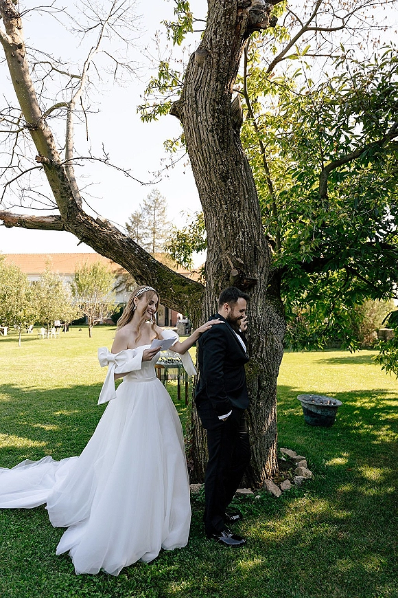 First look moment as bride in wedding dress taps groom in tuxedo on the shoulder while holding vow letters on a garden lawn near a farmhouse