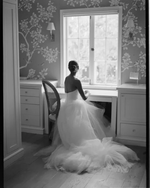 Bridal portrait of a bride from behind in a strapless tulle gown with a long train, seated at a window by a built-in desk chair