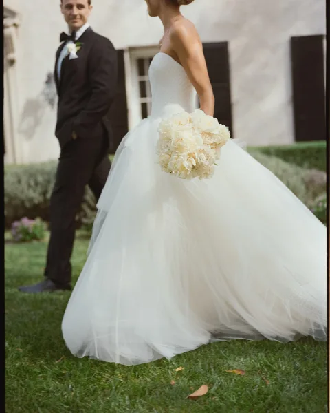 Couple portrait of bride in a strapless tulle gown holding white rose bouquet beside groom in tuxedo by a stucco building and hedges