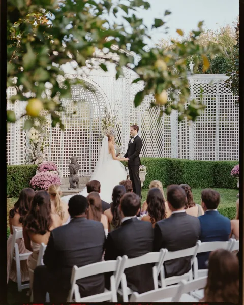 Ceremony moment as bride and groom hold hands at the white lattice altar, veil and tux visible, surrounded by garden flowers and chairs