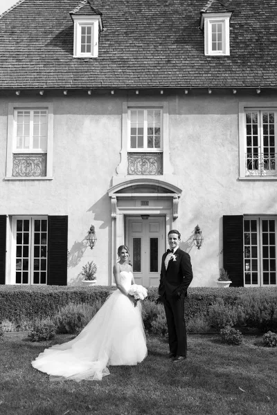Couple portrait in a black and white wedding portrait style, bride holding bouquet beside groom in tuxedo before an estate front door facade