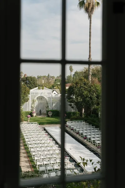 Ceremony setup for an outdoor wedding ceremony with white aisle runner and folding chair rows leading to a lattice altar on a garden lawn
